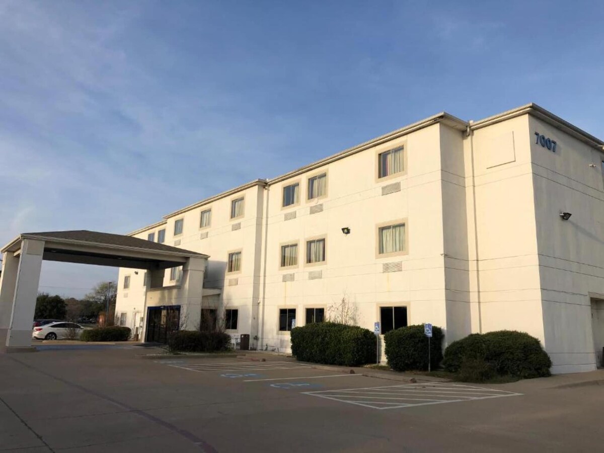 The exterior of a multi-story building is visible, showcasing a simple architectural design with a white façade. Windows are lined symmetrically along the front, and a covered entrance is present. Surrounding shrubbery and a parking area are also depicted.