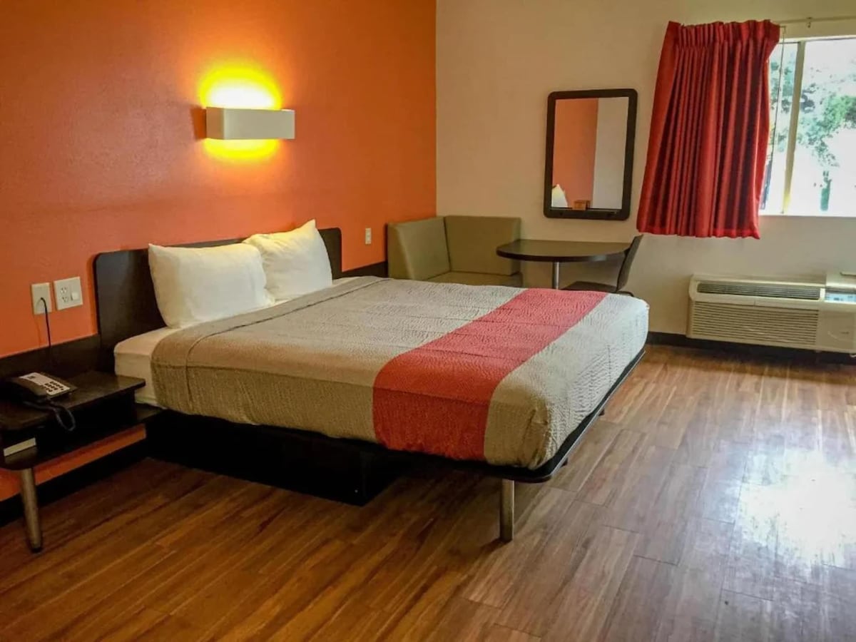 A well-lit room features a spacious bed with neutral-toned bedding, situated against an orange accent wall. A small table and seating area are visible beside the bed. Natural light enters through a window adorned with red curtains. A mirror hangs above a low dresser.