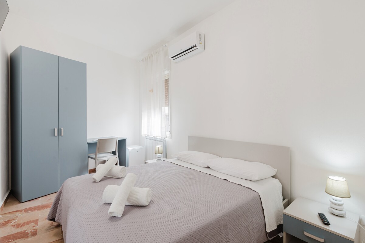 A well-appointed bedroom features a spacious bed with crisp linens, flanked by bedside tables with lamps. A wardrobe provides ample storage, and a desk with a chair offers a dedicated workspace. Natural light filters through the window, enhancing the bright and airy atmosphere.