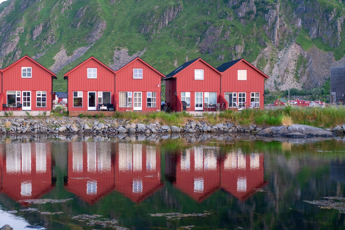 A row of red cabins is positioned along a water's edge, reflecting beautifully on the surface. Green mountains rise in the background, creating a serene landscape. Large windows provide natural light to the interiors, while outdoor seating areas are visible on wooden decks.