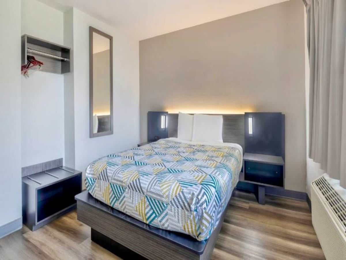 A well-lit bedroom features a spacious bed with a patterned blanket. Nightstands are positioned on either side of the bed, and a mirrored closet reflects the light. A window with a curtain provides natural light, complementing the neutral-toned walls.