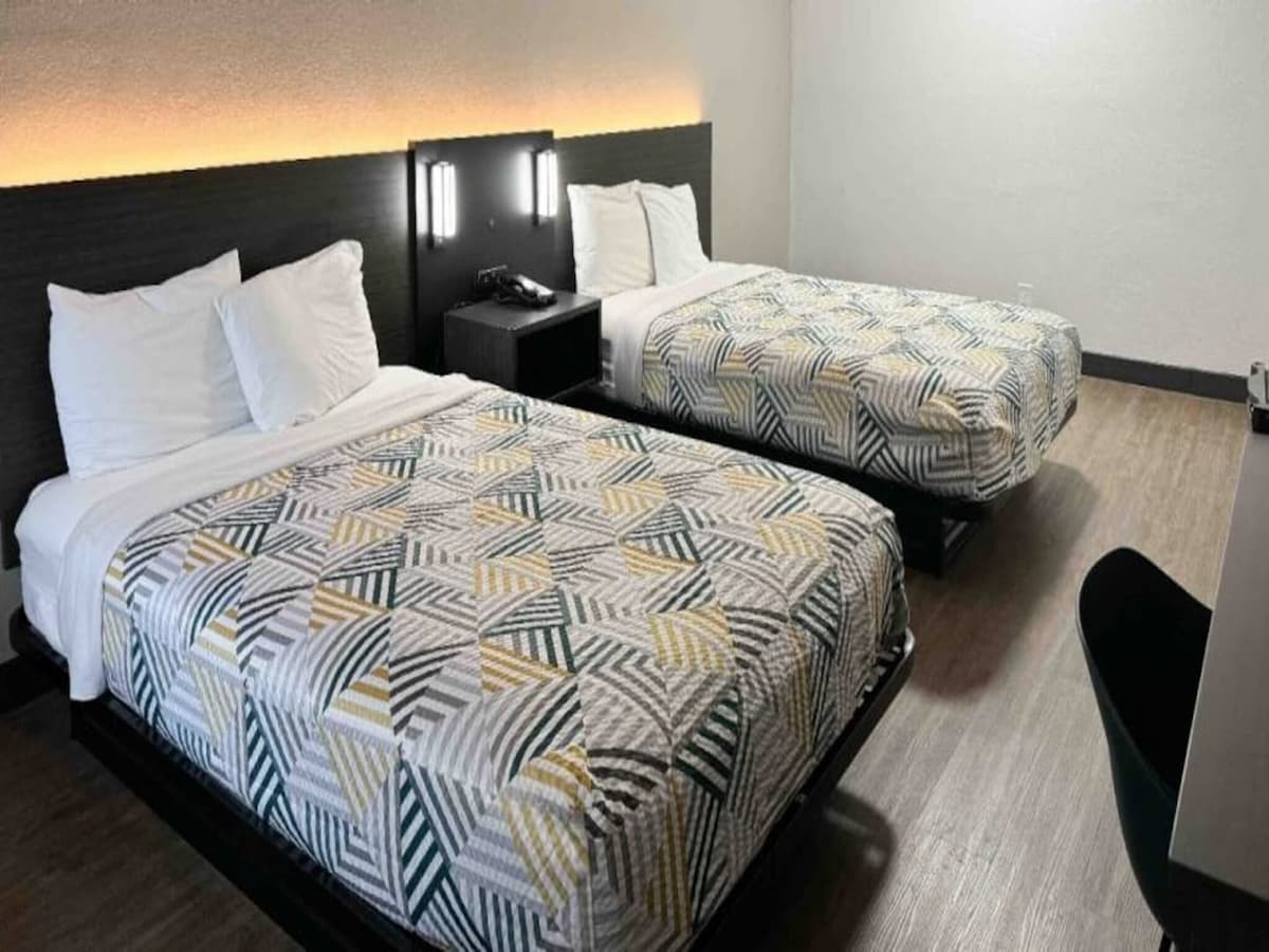 The room features two full beds, each topped with patterned bedding. A bedside table is positioned between the beds, and a phone is visible on the nightstand. Soft lighting illuminates the headboard, while a section of the room shows dark wood flooring.