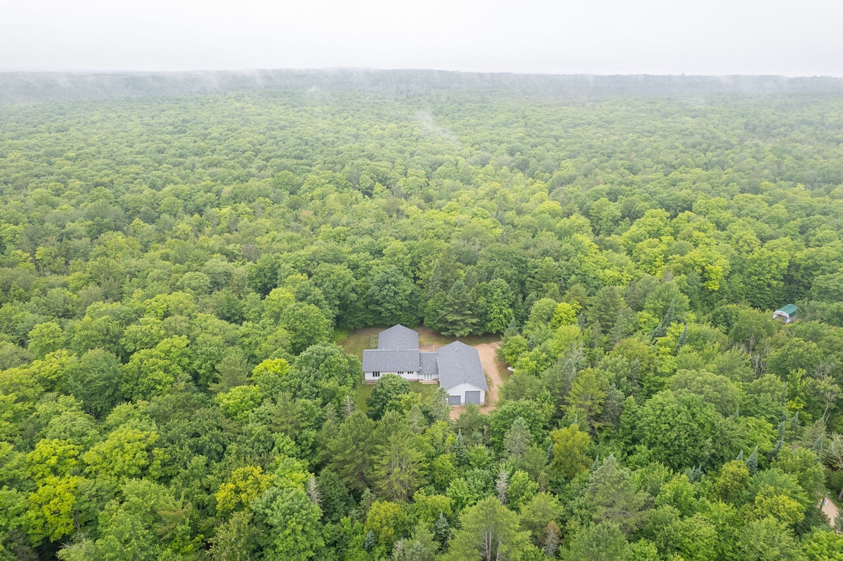 An aerial view captures a spacious retreat nestled within a lush, green forest. The property is surrounded by dense trees, emphasizing its seclusion and natural environment. A winding driveway leads to the home, which blends harmoniously with its wooded surroundings.
