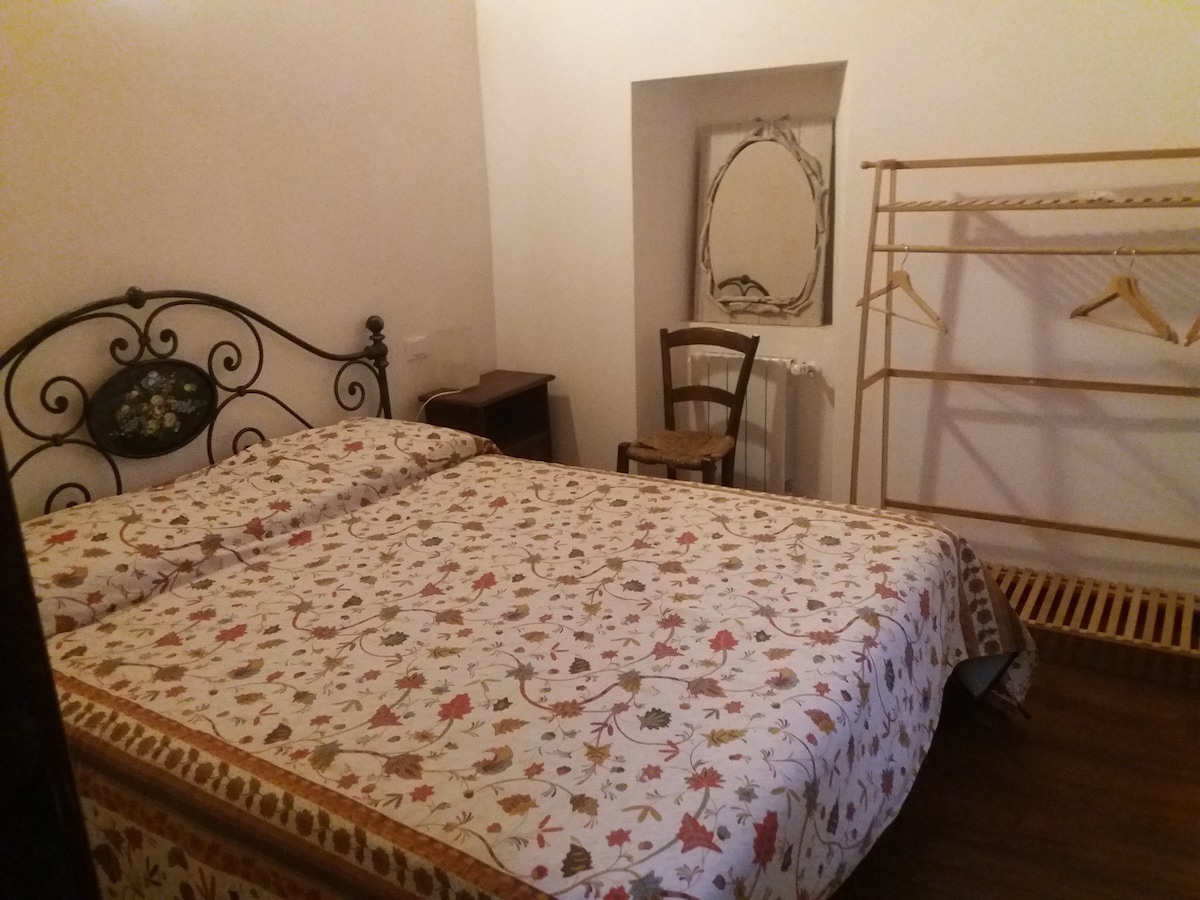 A simple bedroom features two single beds adorned with a floral patterned cover. A mirror hangs above a small wooden nightstand. A wooden chair is positioned nearby. An open clothing rack is available for guests, with natural light illuminating the room.