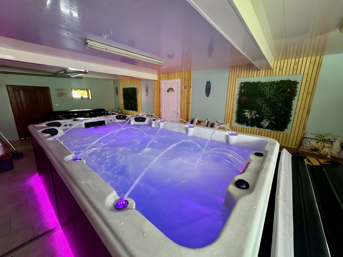 A spacious wellness area features a swim spa with illuminated jet streams, surrounded by a sleek design. Soft purple lighting enhances the atmosphere, while greenery decorates the walls, contributing to a calming environment. Additional wellness facilities are also visible in the space.