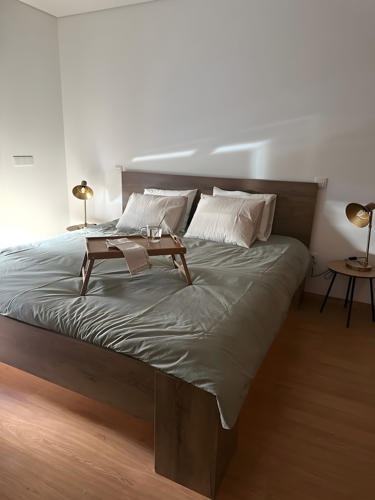 A spacious bed with a sturdy wooden frame is dressed in soft, neutral-toned linens. Two bedside lamps provide gentle lighting, and a breakfast tray rests on the bed, enhancing the cozy setting. Natural light enters through a nearby window, softly illuminating the room.