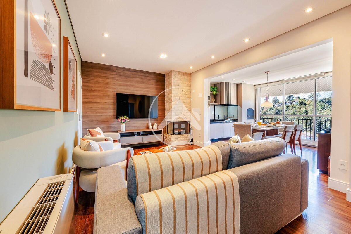 A welcoming living area is highlighted by a comfortable gray sectional sofa with a striped throw. A flat-screen TV is mounted on the wooden accent wall. Light filters in from large windows that open to a view of greenery beyond, enhancing the inviting atmosphere.