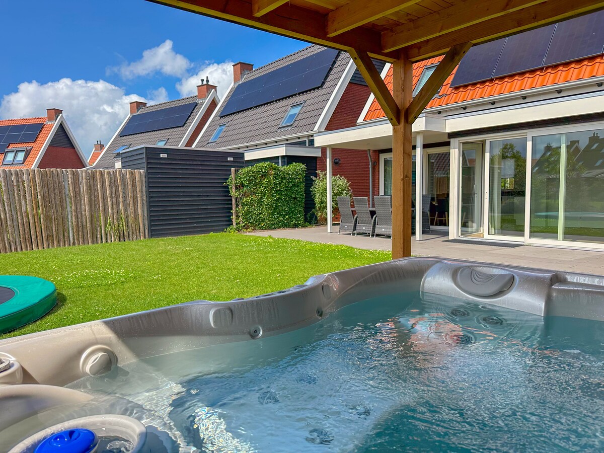 A private garden is featured with well-maintained grass and surrounded by fences. A spa bath is positioned under a wooden pergola, with bubbles gently rising to the surface. In the background, modern houses with solar panels are visible, alongside outdoor seating arrangements.