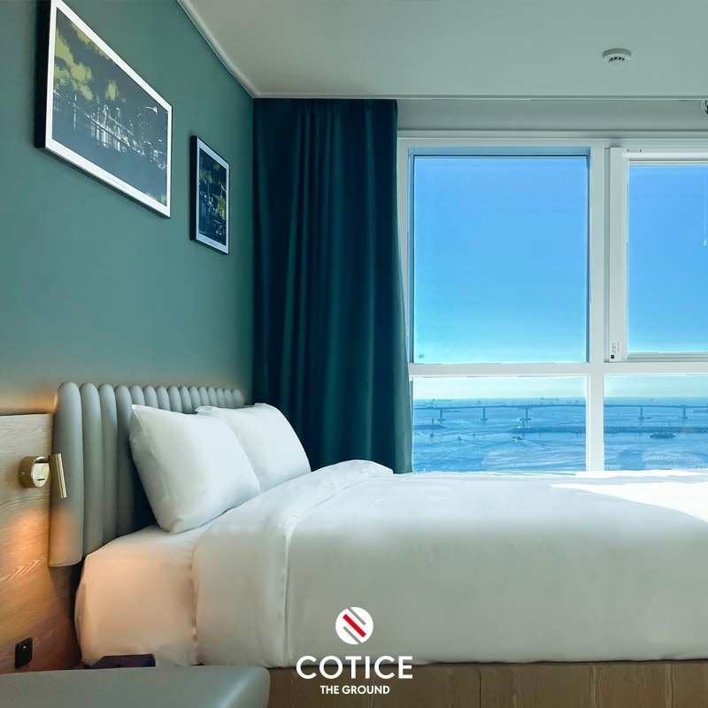 A well-lit sleeping area features a spacious bed with a plush headboard and soft white linens. Colorful artwork adorns the walls, complemented by a large window that offers a clear view of the outside. Soft natural light brightens the room, enhancing its tranquil ambiance.