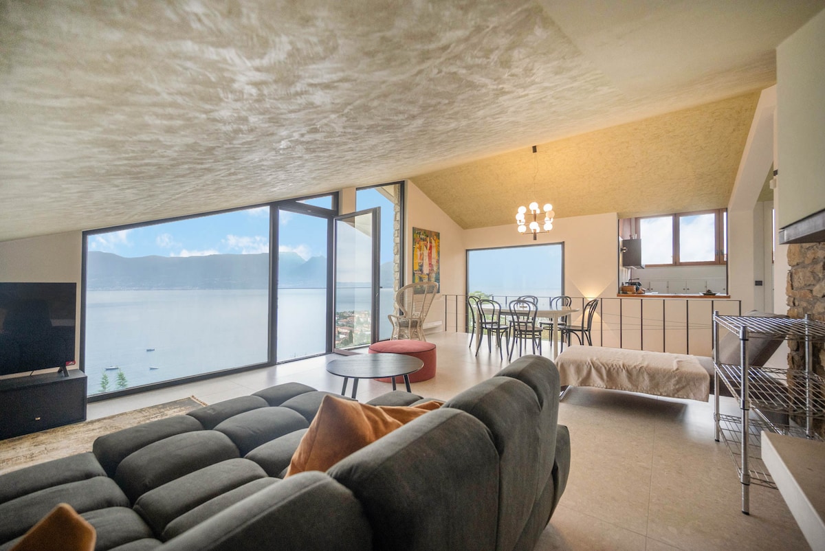 A spacious living area features large glass windows, offering views of a serene lake and distant mountains. Comfortable seating arrangements are visible, along with a dining area that complements the open layout. Neutral tones in the décor enhance the relaxing environment.