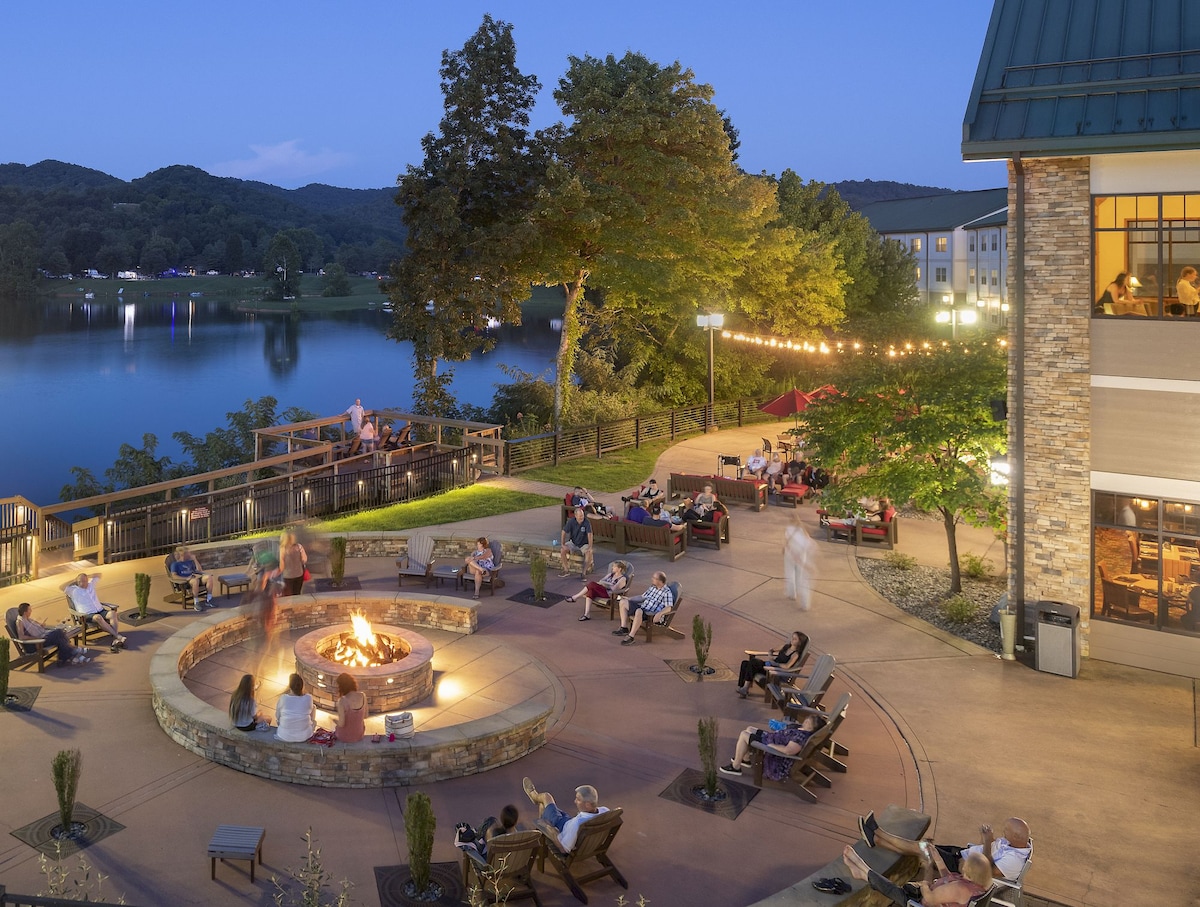 An outdoor fire pit area is surrounded by seating, inviting guests to gather and enjoy the serene lakeside setting. Soft lighting enhances the atmosphere as people relax, with scenic views of the water and wooded mountains in the background under a twilight sky.