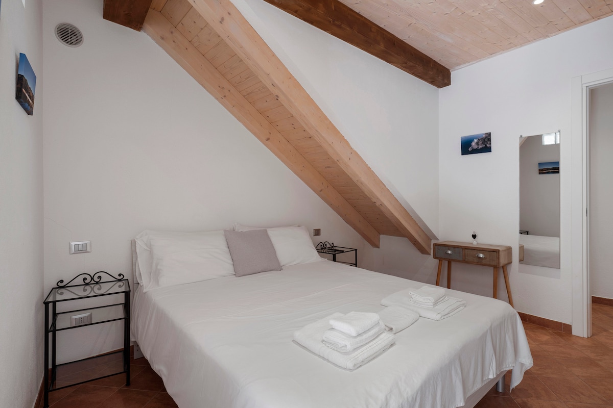 A spacious bedroom features a sloped wooden ceiling, enhancing the room's character. A large bed is neatly arranged with fresh linens and towels. An elegant mirror and a wooden desk add functionality, while a bedside table provides storage. Natural light enters through a nearby window.