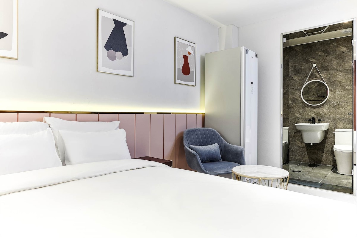 A contemporary room features a spacious bed with crisp white linens and a minimalist design. A blue armchair is positioned near a small round table. A refrigerator and a modern bathroom are visible in the background, complemented by framed art on the walls.