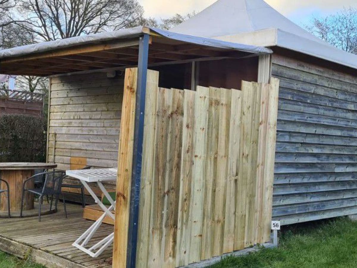 A wooden terrace is displayed, featuring a table and two chairs for outdoor seating. The space is partially enclosed by wooden panels, providing privacy. The structure's exterior is lined with wooden slats, blending with the natural surroundings.