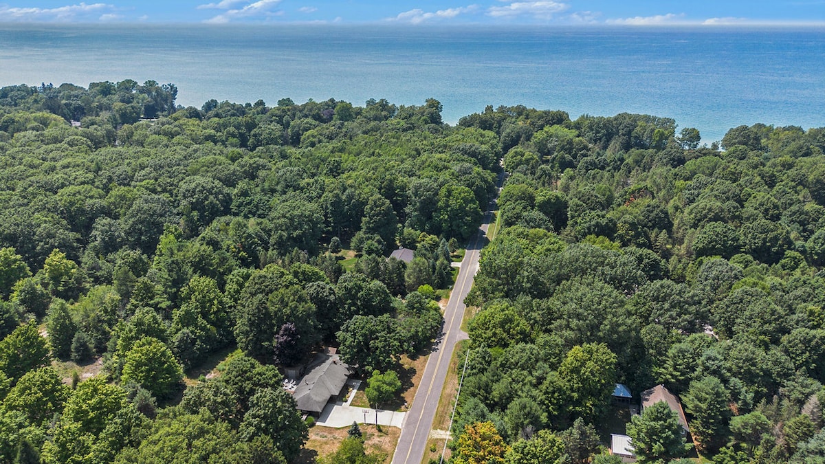 An aerial view showcases a lush green landscape dotted with trees, revealing a winding road leading towards the coastline. The tranquil blue waters of the lake are visible in the distance, surrounded by a canopy of greenery, creating a serene natural setting.