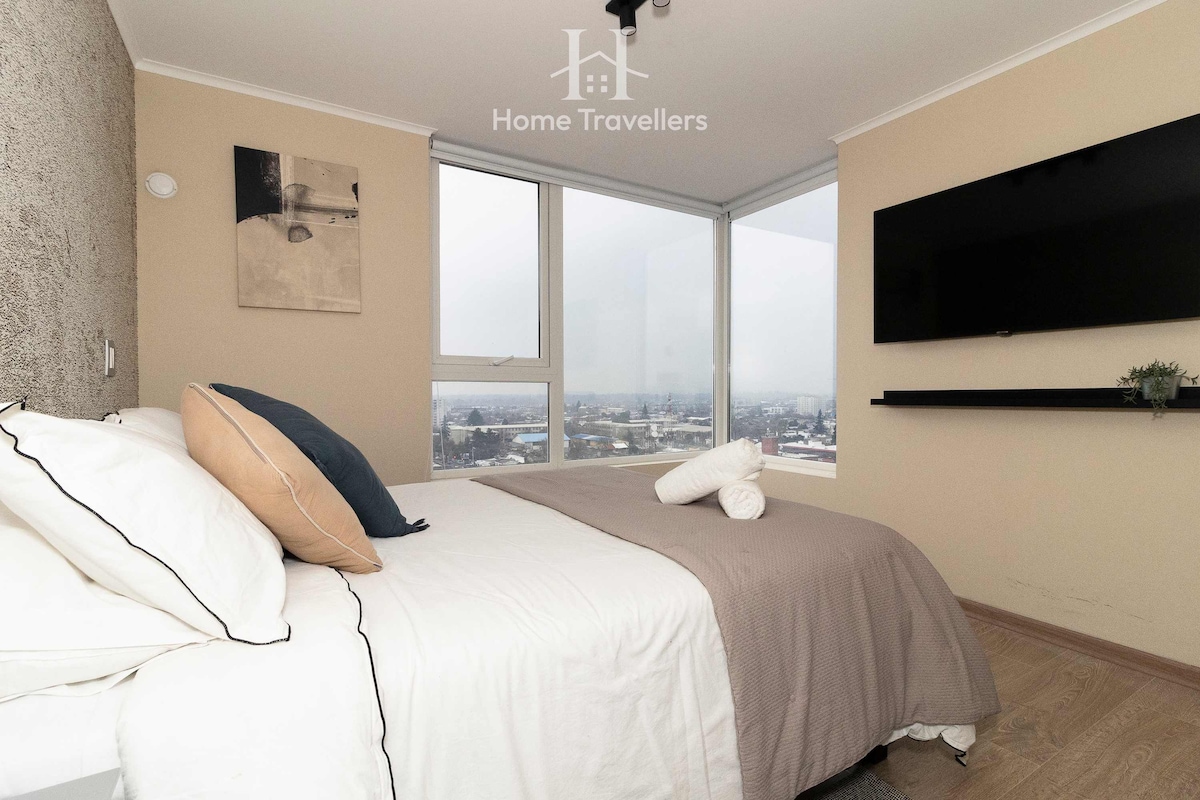 A cozy bedroom features a spacious bed with neutral bedding and accent pillows. Large windows offer expansive views of the surrounding area. A minimalist shelf holds a small plant, and a wall-mounted TV is positioned across from the bed.