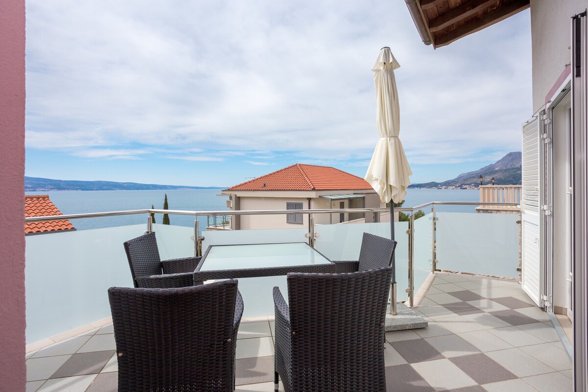 A private terrace is displayed with a glass-topped dining table surrounded by four black wicker chairs. The view includes the sea and nearby rooftops, with a white umbrella positioned for shade against the open sky.