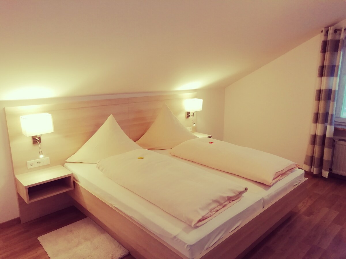 A cozy attic bedroom features a spacious bed adorned with light bedding. Soft lighting from wall sconces creates a gentle glow. Wooden flooring complements the neutral walls, and a small bedside table is positioned beside the bed. A window with light curtains allows natural light to filter in.