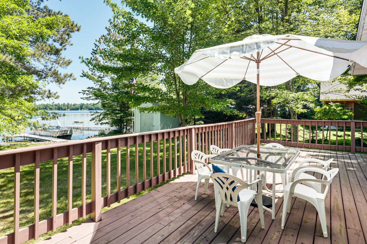 A spacious deck features a glass-top dining table surrounded by white plastic chairs, set against a backdrop of lush greenery and a view of Solberg Lake. A large umbrella provides shade, enhancing the outdoor dining experience in a serene environment.