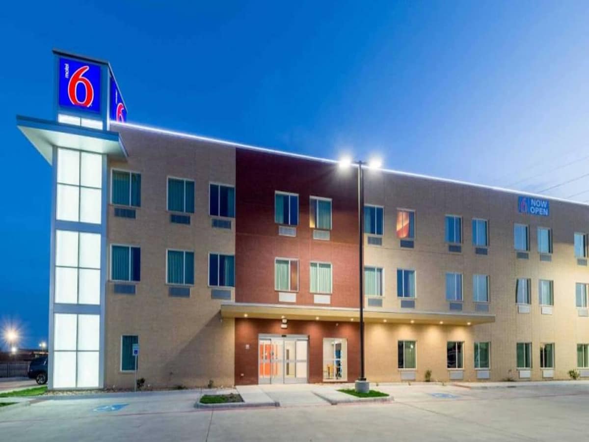 The exterior of the Motel 6 Fort Worth is shown in the evening, featuring a modern façade with a combination of tan and brick colors. The well-lit entrance is flanked by large windows, and the iconic Motel 6 sign stands prominently above.