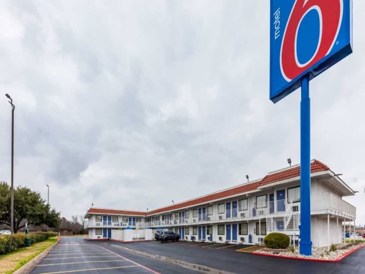 The exterior of Motel 6 is depicted, featuring a two-story building with exterior corridors. The signage displays the brand name prominently. A well-maintained parking area is visible in front, along with manicured landscaping and a cloudy sky in the background.
