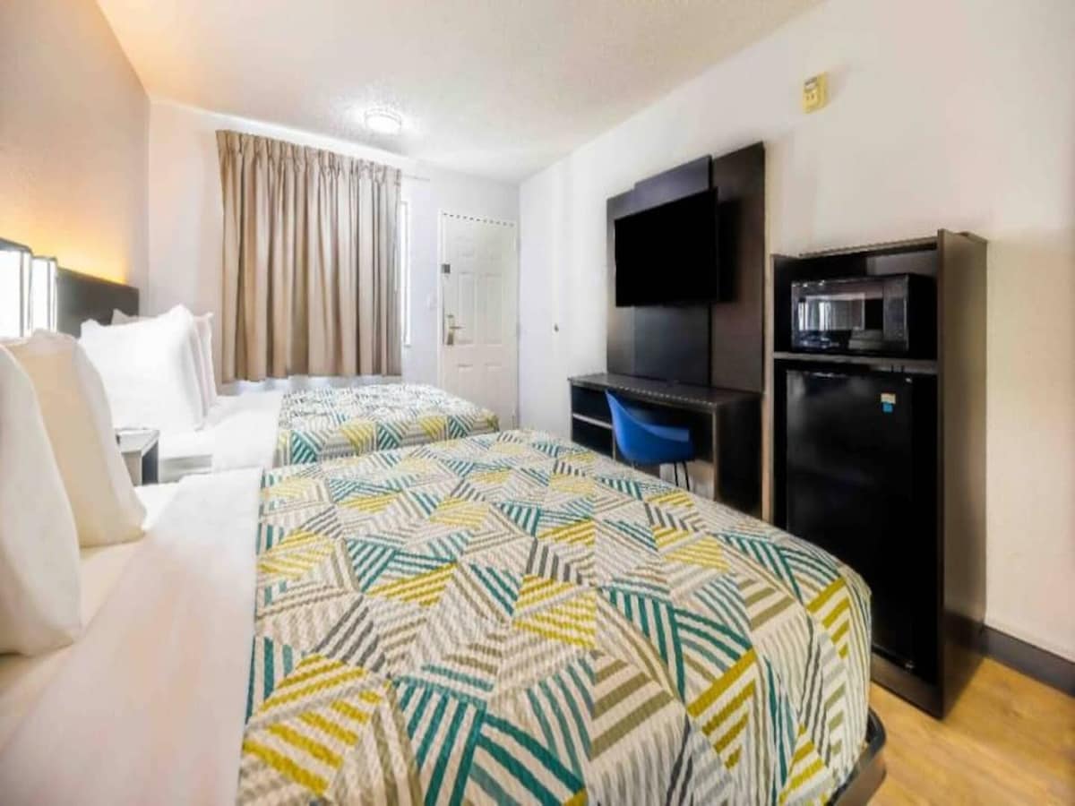 A bright room is equipped with two full beds, adorned with patterned bedspreads. A flat-screen TV is mounted on the wall, and a small desk with a blue chair is present, alongside a microwave and mini-fridge. Natural light filters through the window with drapes.