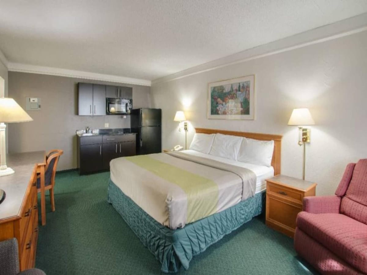 A spacious room features a king-sized bed topped with white linens. A kitchenette with dark cabinetry, a refrigerator, and a microwave is visible. Lighting fixtures on either side of the bed provide soft illumination. A comfortable armchair and a desk are included within the area.