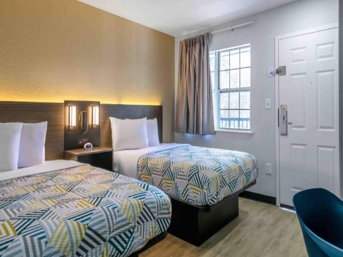 A well-lit guest room features two queen beds, each adorned with patterned bedspreads. Nightstands with lamps are positioned above the beds. A window with sheer curtains allows natural light, and a chair is situated near the entrance door.