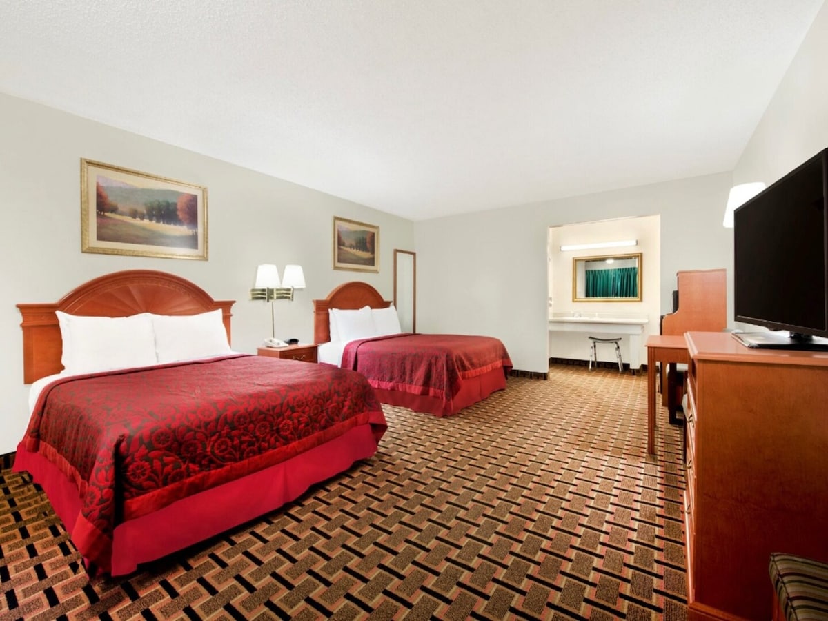 The spacious guest room features two queen beds with dark red bedding. A decorative painting hangs above each bed. A wooden desk and chair are positioned near a wall-mounted television. Soft carpeting adds warmth to the room, while natural light enters through a nearby window.