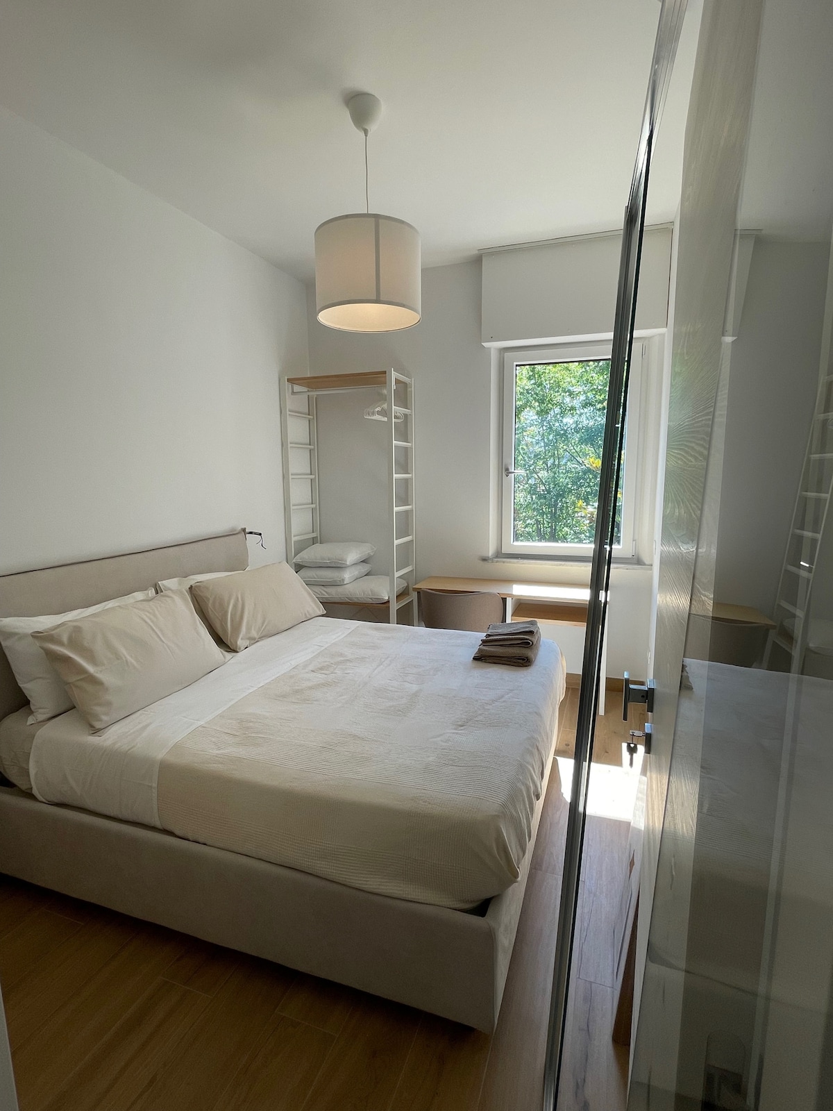 A cozy double room is presented with a spacious bed dressed in neutral linens. Natural light filters through the window, illuminating simple furniture including a desk and shelving unit. Greenery is visible outside, enhancing the tranquil atmosphere.