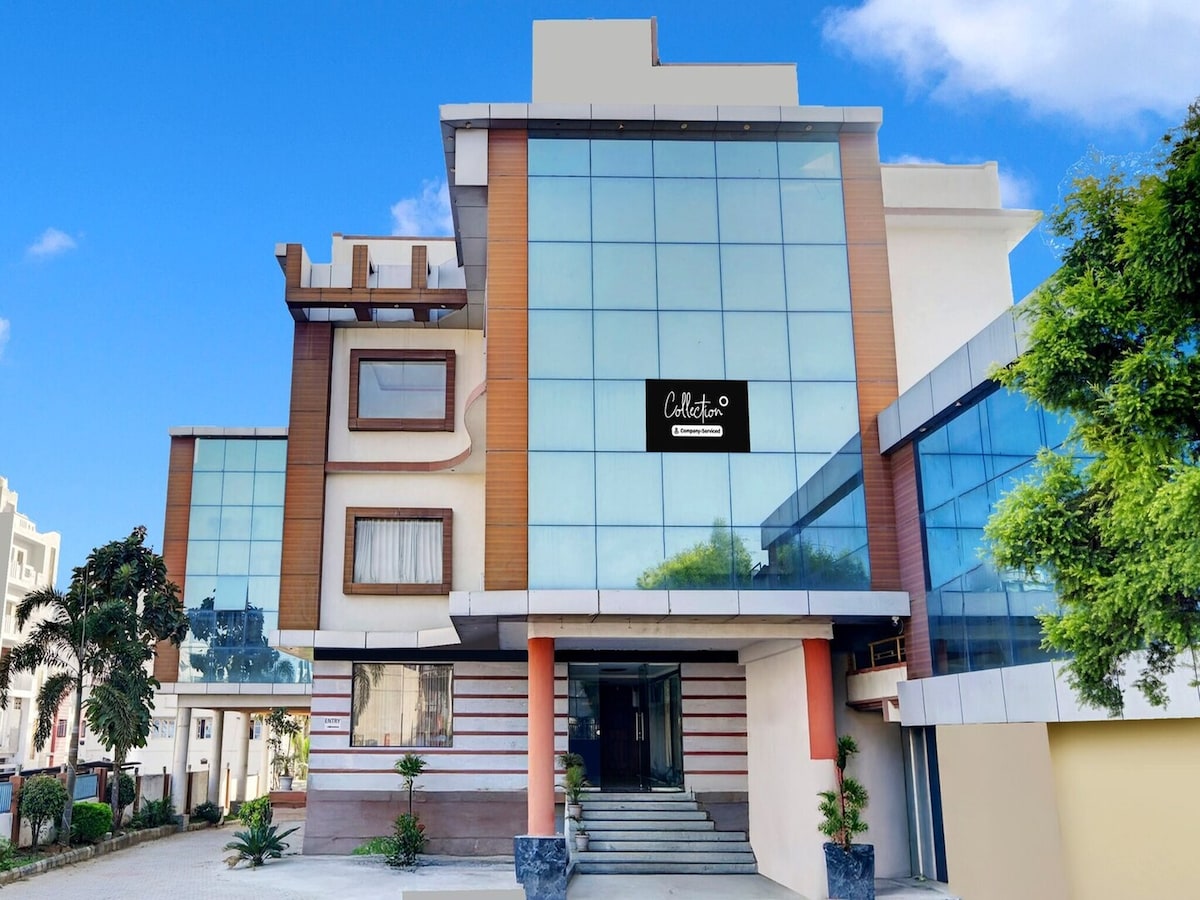 The exterior of the hotel is characterized by a modern design with expansive glass windows, allowing ample natural light. A landscaped entrance is visible, featuring potted plants and a staircase leading to the main entrance, framed by a contemporary facade.