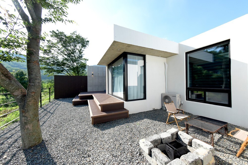 An outdoor area of the villa is presented, featuring wooden decking and a seating arrangement. A fire pit is positioned within a stone border, complemented by gravel flooring. Large windows allow ample natural light, and greenery is visible in the background, enhancing the serene environment.