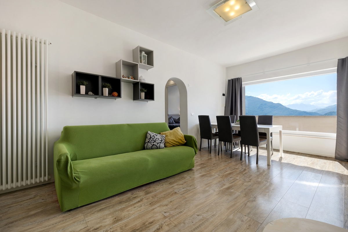 A bright living area features a green sofa accompanied by decorative cushions. A dining table with chairs is positioned near a large window, offering views of the surrounding mountains. Wall shelves display various items, and natural light fills the space through darker curtains.