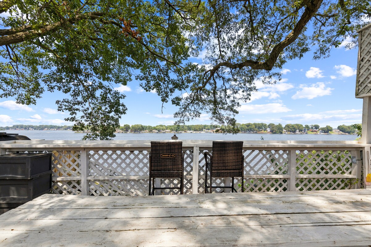 A lakeside deck is presented, featuring a weathered wooden table and two woven chairs positioned for river views. Dappled sunlight filters through the branches of a large tree overhead, casting natural patterns on the deck surface. The expanse of Lake Shafer stretches into the distance.