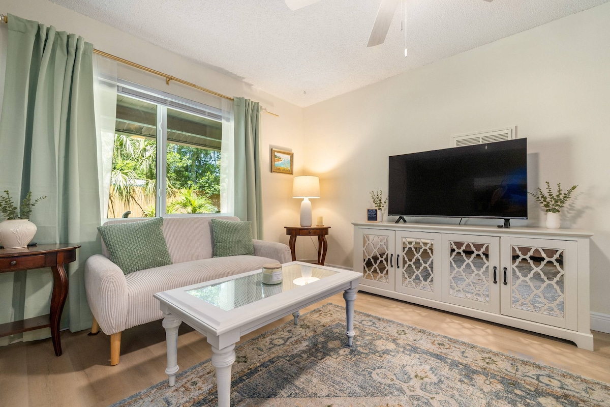 A comfortable living area features a light-colored sofa adorned with decorative cushions, complemented by a white coffee table. Natural light flows through a large window framed by soft green curtains. A flat-screen TV is positioned on a stylish sideboard, enhancing the space.