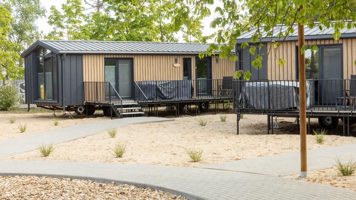 Two modern mobile homes are positioned on raised platforms, surrounded by sandy areas and greenery. Each home features a deck with seating and large windows, providing natural light. A curved pathway of gray pavers leads through the landscape, enhancing accessibility.