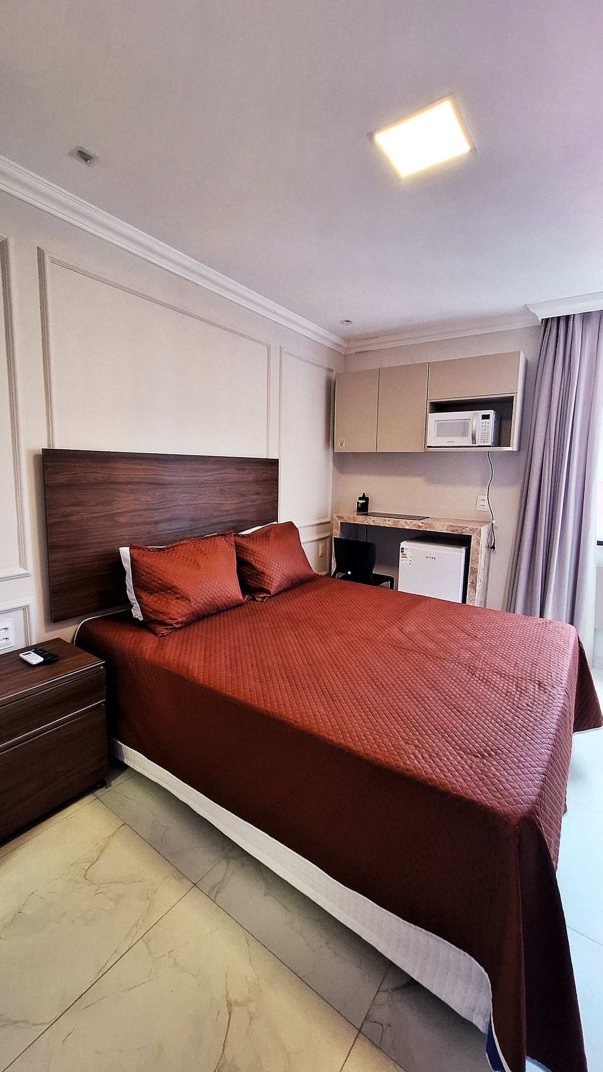 A spacious studio room is equipped with a comfortable bed featuring a textured brown coverlet. A kitchenette area includes a small refrigerator and microwave, while natural light filters in through the curtains beside the window.