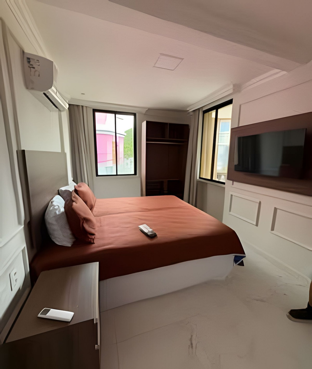A studio room is presented, featuring a spacious bed with a brown coverlet and coordinated pillows. Natural light filters through large windows adorned with light curtains. A flat-screen television is mounted on the wall, and a wardrobe is positioned nearby, complementing the modern decor.