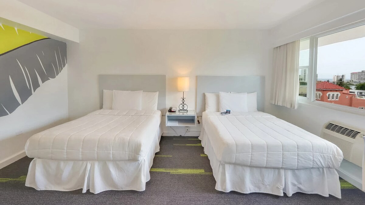 A well-lit room features two spacious beds dressed in white linens, positioned next to a bedside lamp on a nightstand. A window offers natural light, revealing a view of the surrounding area and a muted, contemporary design on the wall.