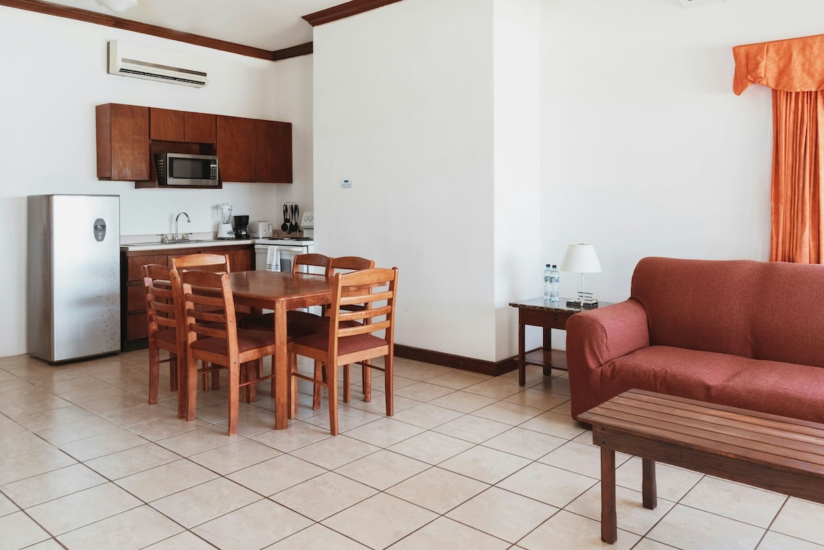 A straightforward living area features a dining table with six wooden chairs and a comfortable sofa. The kitchen includes stainless steel appliances and light wood cabinetry, while a coffee table is positioned in the center. Light beige tiles cover the floor, contributing to an airy feel.