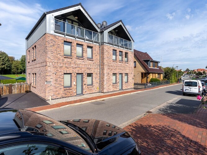 A modern two-story building is depicted, featuring a distinctive brick façade and large windows. The structure is flanked by a neatly paved street and landscaped areas, with green lawns visible in the background. A vehicle is partially seen in the foreground.