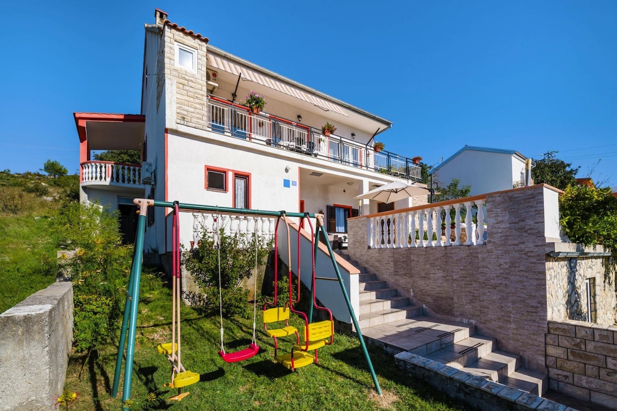 A two-story house is surrounded by greenery, featuring a red and yellow swing set in the foreground. A staircase leads up to the entrance, while a balcony with potted plants extends along the upper level, offering outdoor space for relaxation.