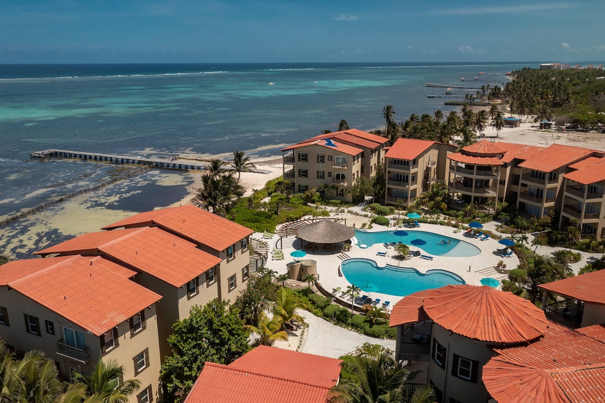 Aerial view of the condominium resort shows multiple buildings with terracotta roofs surrounding a large swimming pool. Lush landscaping leads to the beach, with the turquoise Caribbean Sea extending toward the horizon. A pier is visible in the distance, along with gentle waves.