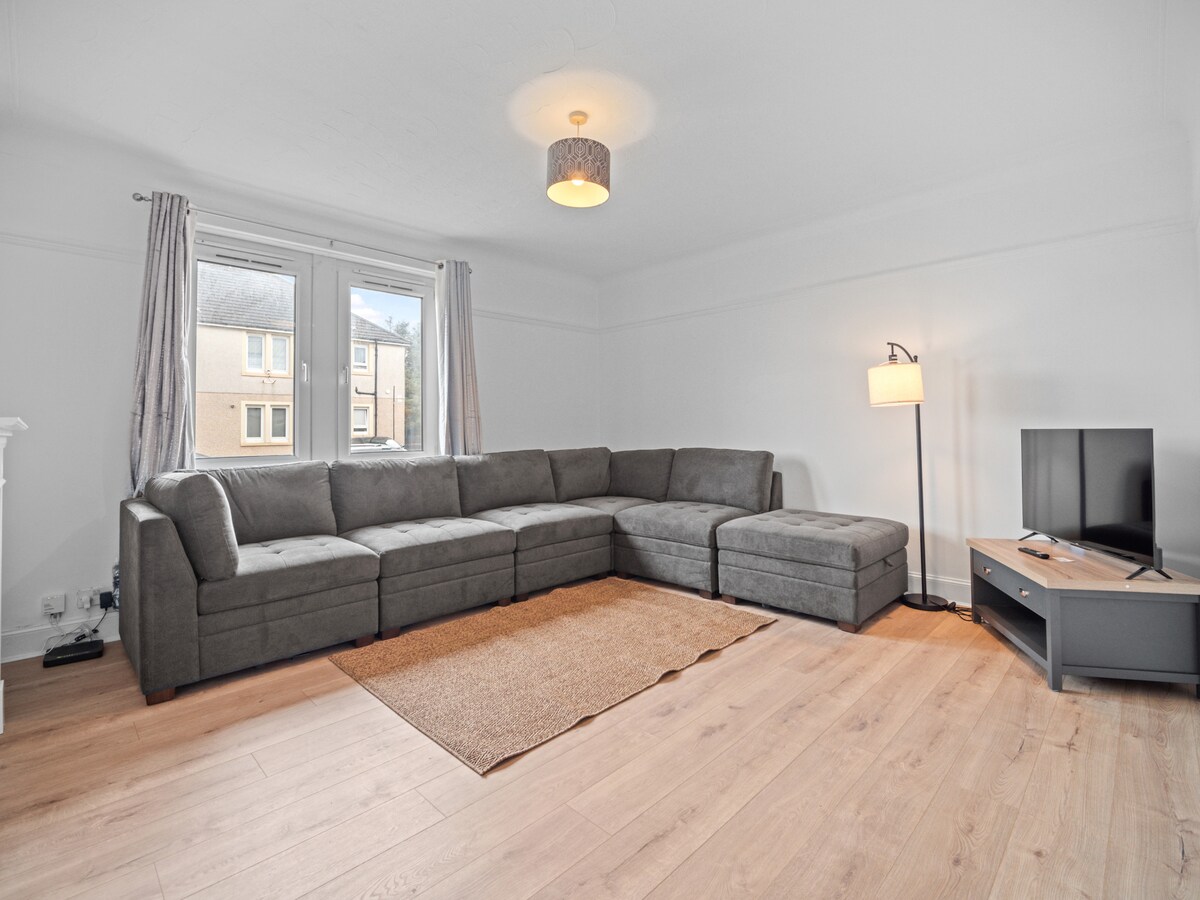 A spacious living area features a large sectional sofa and a light-toned area rug on wooden flooring. Natural light fills the room through two windows, complemented by gray curtains. A simple floor lamp and a flat-screen television are also present.