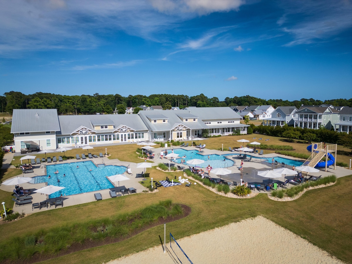 An aerial view captures the expansive pool area, dotted with umbrellas and lounge chairs. Two large pools are visible, surrounded by a well-maintained lawn and a sandy beach area. Nearby, the quaint clubhouse features multiple windows and outdoor seating.