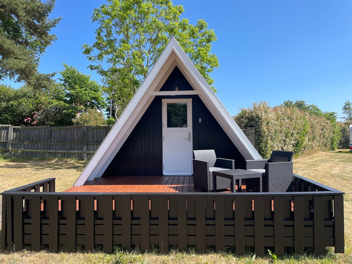 An A-frame cabin is set in a grassy area, featuring a wooden deck with two black wicker chairs. The cabin's front is characterized by a white door and large windows, complemented by a surrounding black wooden fence. Lush greenery is visible in the background.