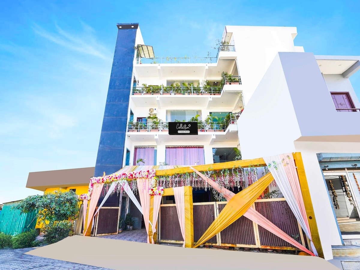 The exterior of the hotel displays a modern design with multiple levels and balconies adorned with greenery. A welcoming entrance is framed by decorative drapes and flowers. The bright blue sky serves as a backdrop, enhancing the building's inviting atmosphere.