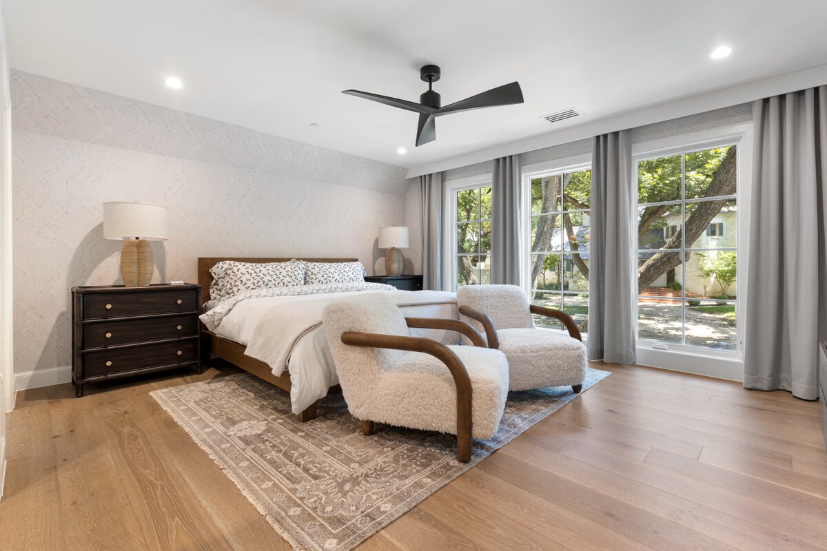 A serene bedroom retreat featuring a king bed framed by textured walls, plush seating, and soft natural light through expansive windows. Layered rugs and warm wood tones create a refined yet inviting space, perfect for restful nights.
