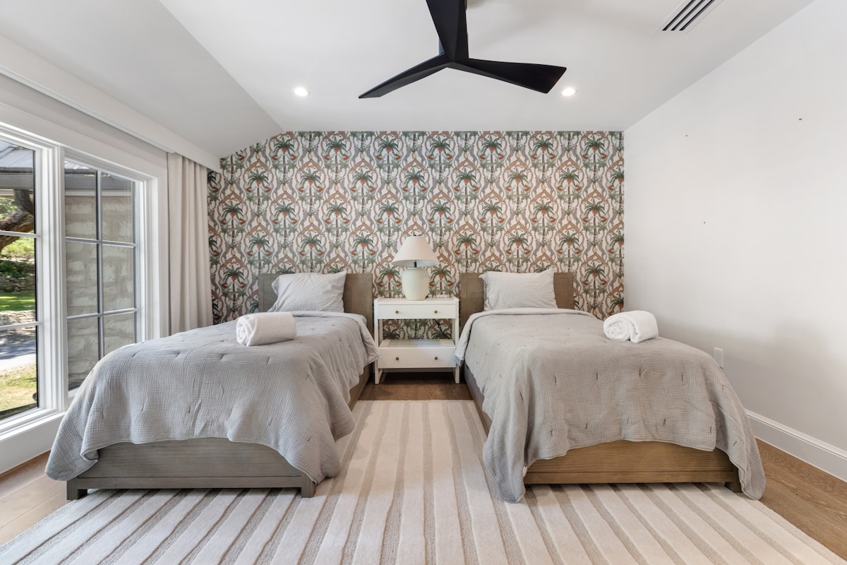This playful bedroom offers two twin beds framed by a bold patterned accent wall, softened with light grey bedding and a striped area rug. Large windows flood the room with natural light, creating a cheerful retreat with easy access to an en suite ba