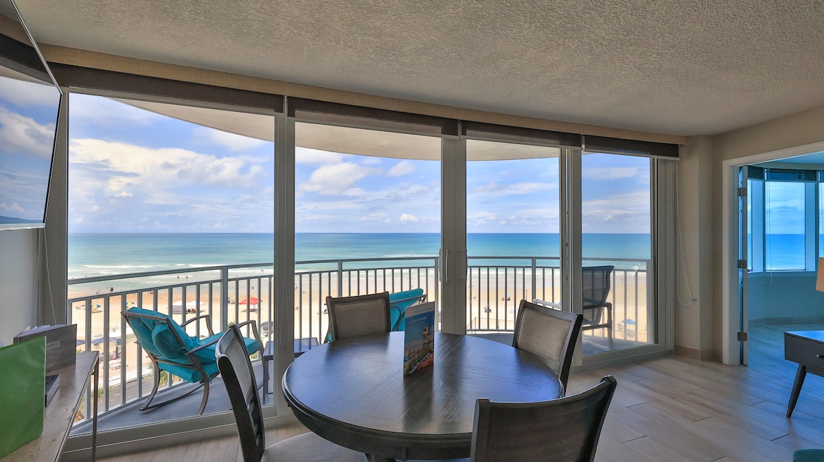 A spacious dining area is highlighted by a round table surrounded by comfortable chairs. Floor-to-ceiling windows offer expansive views of the ocean and beach, with turquoise waters visible under a partly cloudy sky. The design creates a bright and airy atmosphere.
